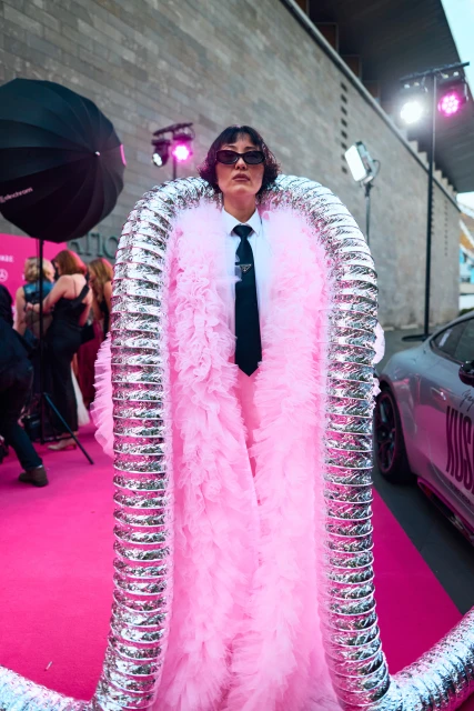 Person on a pink carpet wearing a pink outfit and metallic tubes around them, with a luxury car and event lighting nearby.